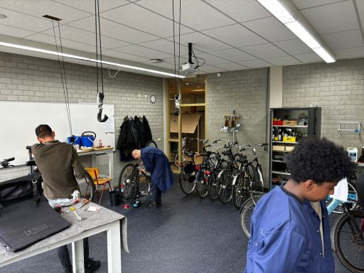 Fietsreparatie in een werkplaats met drie mensen en meerdere fietsen in de achtergrond.
