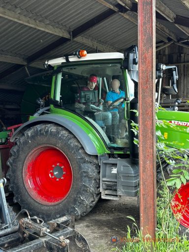 Twee personen zitten in een groene tractor in een schuur.
